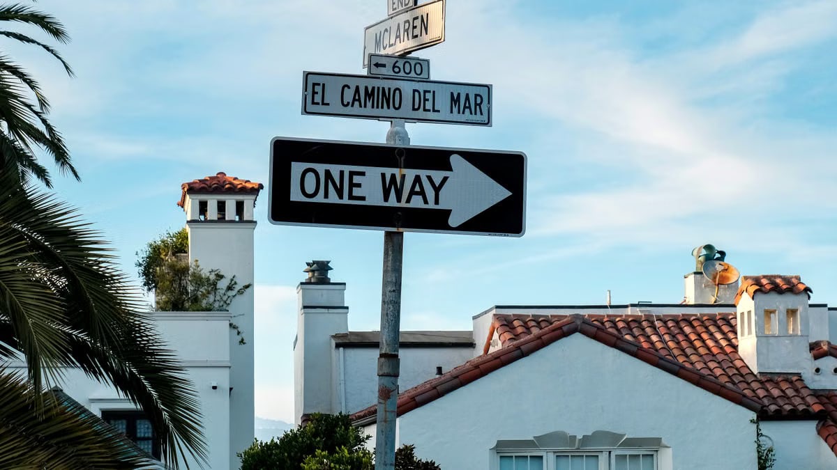 Street signs with Spanish-style architecture, depicting direction and local street names on a sunny day.